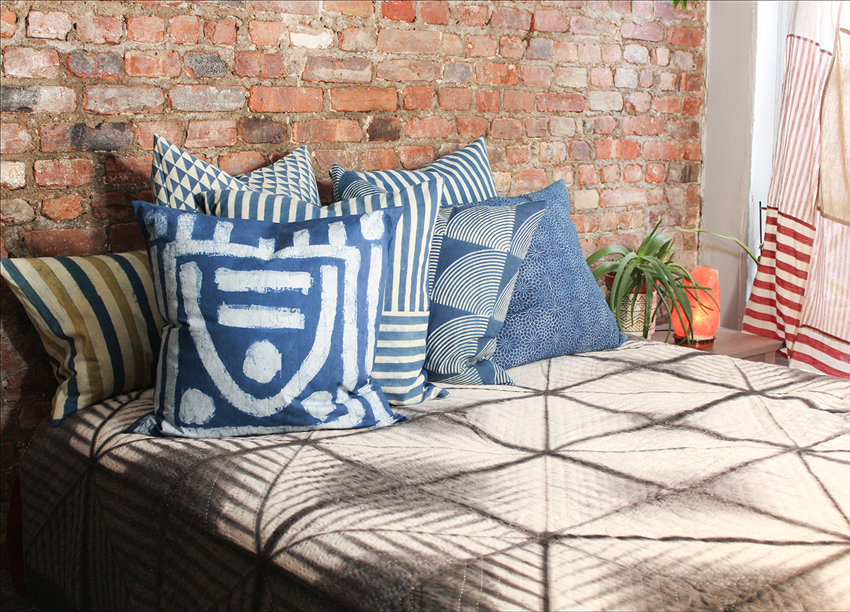 A selection of indigo dyed pillows on a bed with a gray quilt and an exposed brick wall